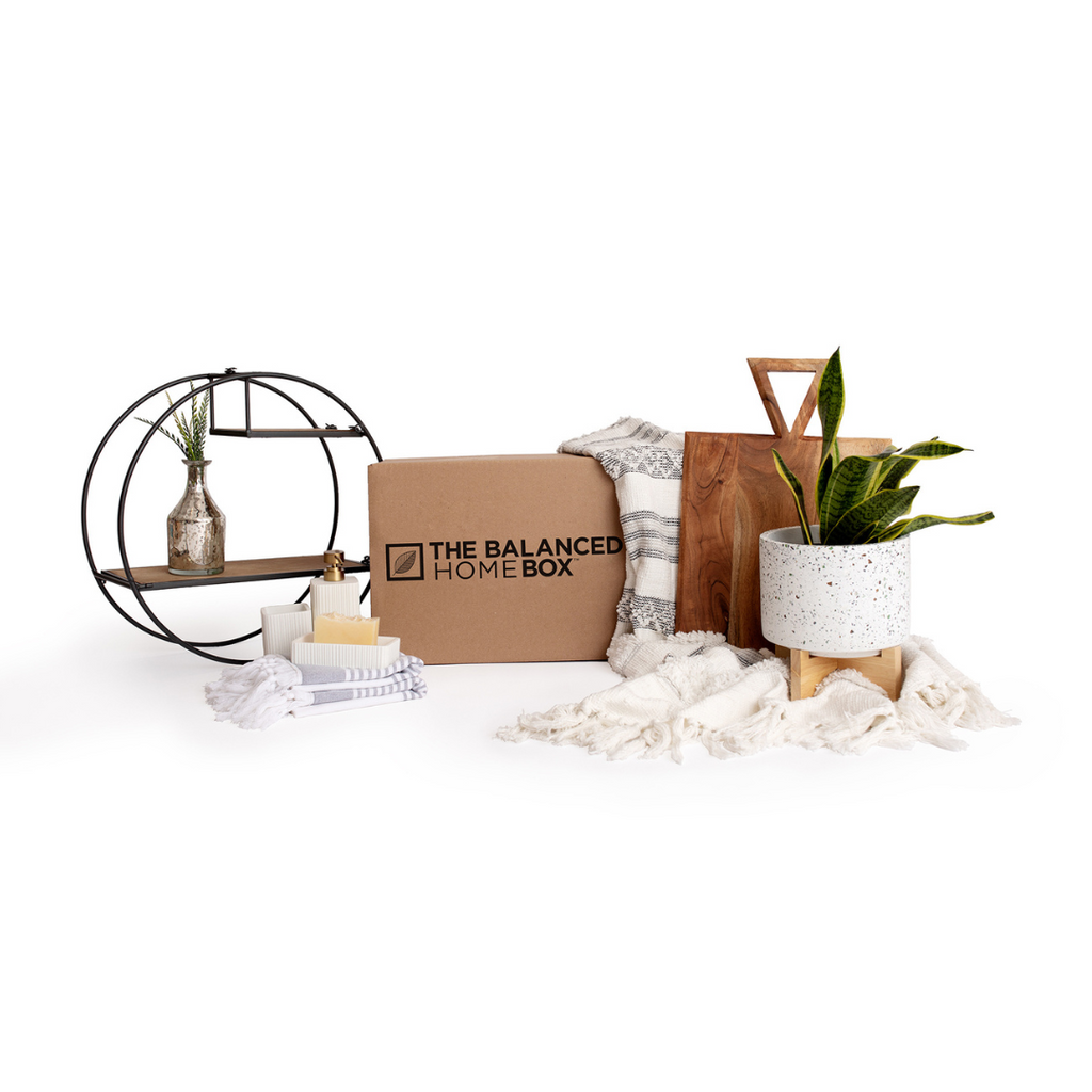 The Balanced Home Box pictured with a circular iron and wood shelf, striped towels with tassels, a luxury soap and toothbrush holder set, a boho throw blanket with tassels, a wood serving tray with a unique triangular handle, and a plant in a white pot with black speckles.