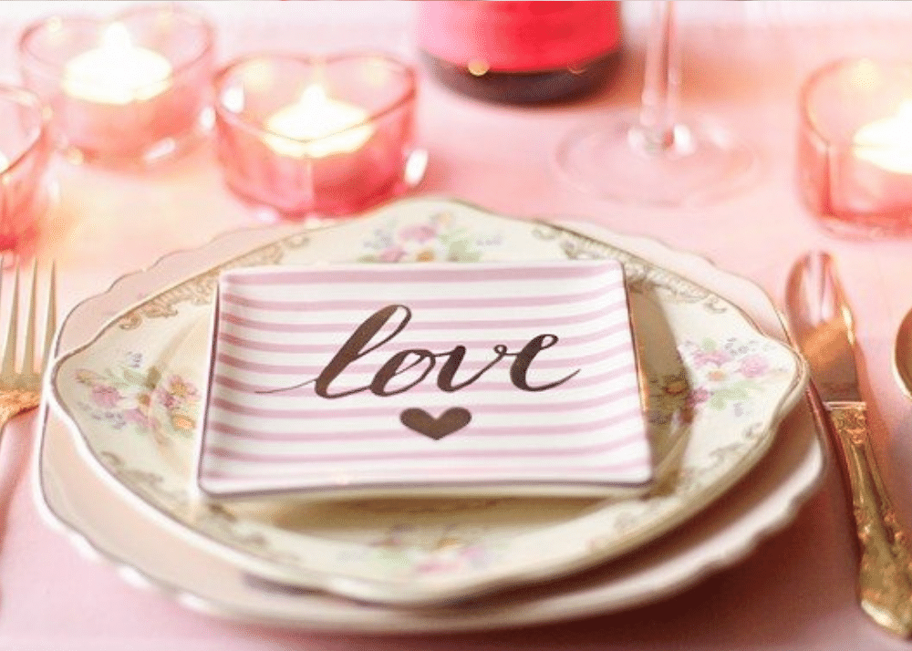 Valentine's Day table setting featuring pink, heart-shaped candle votives with tealight candles and a stack of pretty dishware topped with a smaller 'LOVE' plate with gold cutlery on either side.
