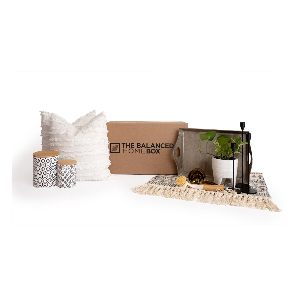 The Balanced Home Box pictured with two plush pillows with white, fringe pillow covers, a pair of trendy cylinder storage containers, a serving tray, a potted plant in a white planter, a small area rug with tassels and a pair of black iron candle holders.