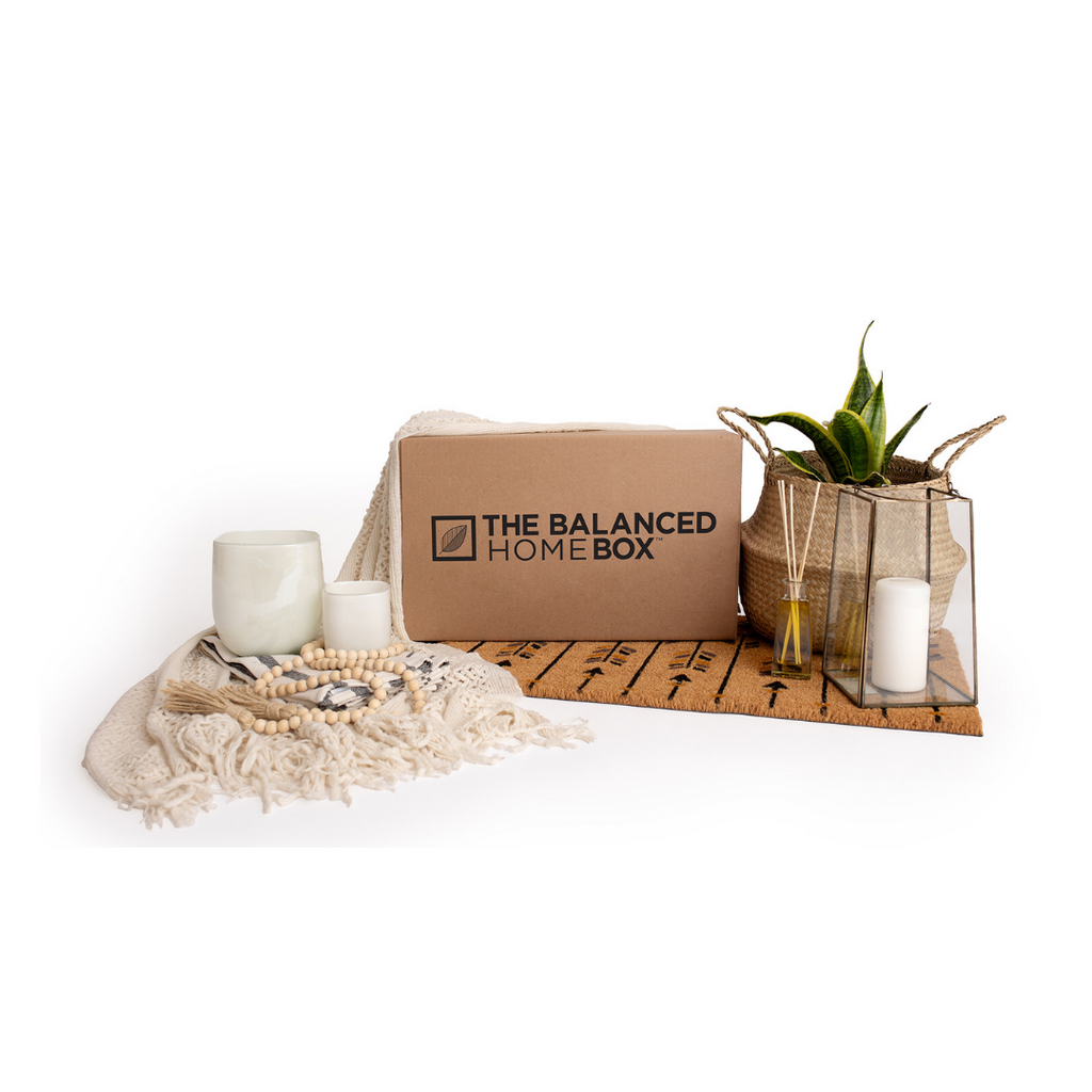 The Balanced Home Box pictured with a pair of white planters, a cream-coloured throw blanket with tassels, a wicker basket with a plant inside, a large glass candle holder with a white cylinder candle inside and an essential oil reed diffuser with sticks inside. All sitting on top of a door mat with a black arrow pattern.