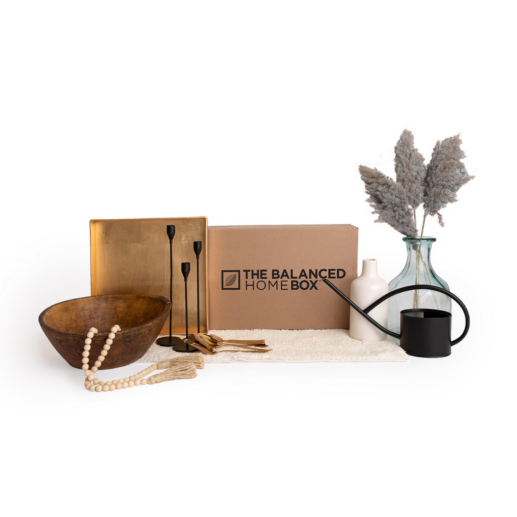 The Balanced Home Box pictured with a gold, square tray, a large wooden bowl, a rustic wood bead garland, a set of three black iron candle stick holders, a set of gold cutlery, a unique white vase, a clear vase and a trendy black watering can. All sitting on a fluffy white bath mat.